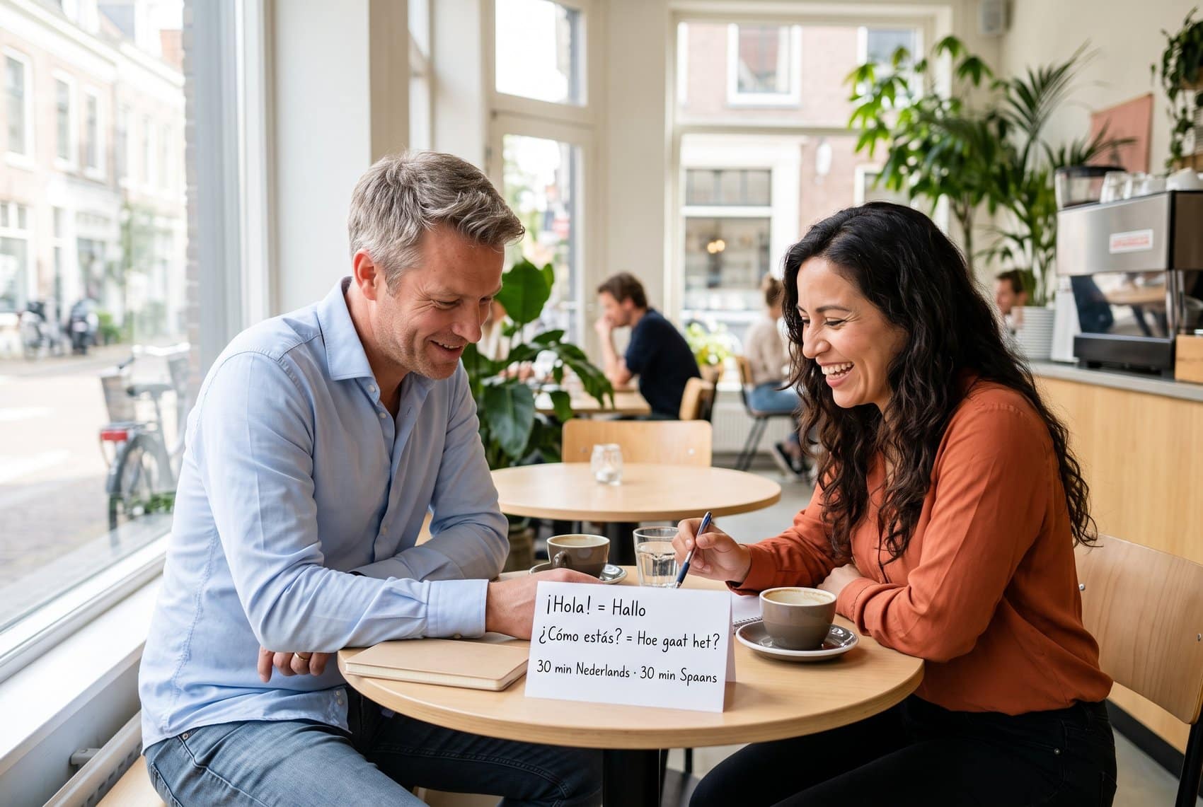 Taaltandem in een café: een Nederlandse man en een Spaanstalige vrouw oefenen samen, met een kaartje met ¡Hola! = Hallo en 30 minuten Nederlands, 30 minuten Spaans